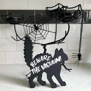 May include: Black cat silhouette with the text "Beware the Vacuum!" on a white background. The cat is standing on a black metal spider web candle holder with three candle holders.