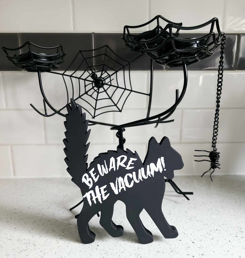 May include: Black cat silhouette with the text "Beware the Vacuum!" on a white background. The cat is standing on a black metal spider web candle holder with three candle holders.