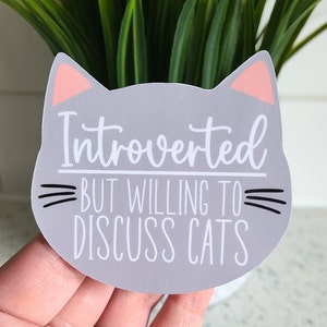 May include: Grey cat-shaped sticker with pink ears and black whiskers. The sticker says "Introverted but willing to discuss cats".