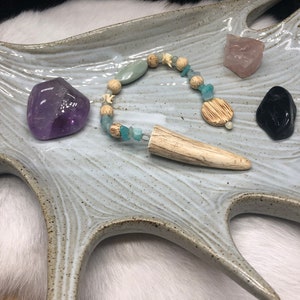 May include: A white ceramic antler-shaped dish with a beaded necklace, a purple amethyst crystal, a pink rose quartz crystal, and a black obsidian crystal.