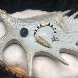 May include: A white ceramic antler dish with a green stone, a bone pendant with blue beads, a clear quartz crystal, and a silver snowflake charm.