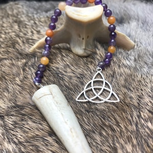 May include: A white bone pendulum with a silver Celtic knot pendant hanging from a beaded necklace with purple, orange, and brown beads.