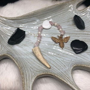 Puede incluir: Un plato de cerámica blanca con un esmalte gris claro presenta un collar de cuentas rosadas y blancas con un colgante de hueso, un dije de abeja dorada y tres piedras negras.