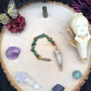 May include: A green and brown beaded necklace with a gold pendant and a white antler charm. The necklace is displayed on a wooden surface with various crystals and a butterfly.