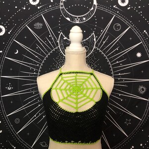 May include: A black crochet top with a green spiderweb design. The top is on a mannequin against a black background with white stars and moons.