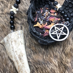May include: A black beaded necklace with a pentagram charm hangs from a white antler point. The necklace is resting in a black, winged bowl filled with dried herbs and flowers.