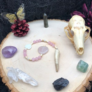 Puede incluir: Un círculo de madera con una variedad de cristales, una pequeña calavera de animal, una mariposa, una piña y un collar de cuentas rosadas con un colgante de hueso.