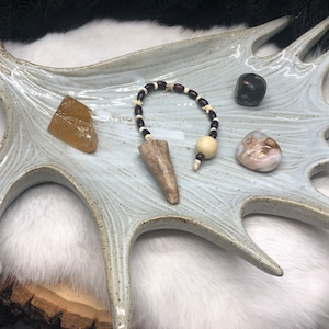 May include: A white ceramic antler-shaped dish with a brown and white beaded pendulum, a brown stone, a clear yellow stone, and a white and brown stone.