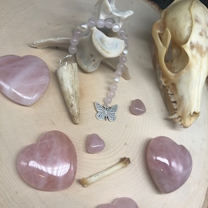 May include: A pink rose quartz crystal heart charm hangs from a silver chain with a butterfly pendant. The charm is surrounded by a strand of pink rose quartz beads and a bone pendant. The background is a wooden surface with a bone skull and other rose quartz crystals.