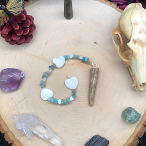 May include: A wooden tray with a variety of crystals, a butterfly, a pine cone, a skull, and a necklace with white heart charms and blue and green stones.