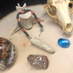 May include: A collection of crystals, a seashell, a bone pendant, and a skull on a wooden surface. The crystals are a clear quartz, a blue crystal, and a red crystal. The bone pendant is attached to a black beaded necklace with red beads.