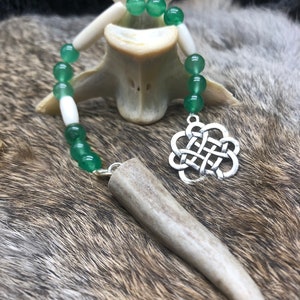 May include: A green and white beaded necklace with a silver Celtic knot pendant and a long antler charm. The necklace is displayed on a white antler.
