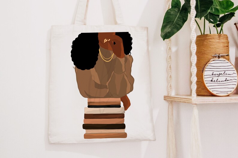 Puede incluir: Una bolsa de tela blanca con una ilustraci&oacute;n marr&oacute;n y negra de una mujer con el pelo negro rizado que lleva un jersey marr&oacute;n y joyas de oro. La mujer est&aacute; sentada sobre una pila de libros.