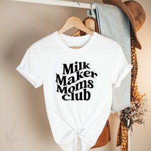 Puede incluir: Camiseta blanca con texto negro que dice "Milk Maker Moms Club" en una fuente retro.