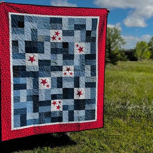 May include: A handmade quilt with a patchwork design in shades of blue and white, accented with red stars. The quilt has a red border with a white inner border. The quilt is displayed outdoors against a backdrop of green grass and a blue sky.