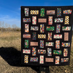 May include: A black quilt with a repeating pattern of rectangular blocks featuring images of darts, pool balls, beer mugs, and other bar-related items. The quilt is displayed outdoors against a backdrop of a blue sky and dry grass.