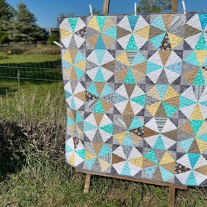 May include: A handmade quilt with a geometric pattern of triangles in shades of gray, teal, yellow, and brown. The quilt is displayed outdoors on a wooden easel, showcasing its intricate design and craftsmanship. The quilt is approximately 60 inches by 70 inches.