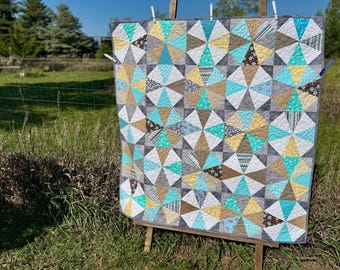 May include: A handmade quilt with a geometric pattern of triangles in shades of gray, teal, yellow, and brown. The quilt is displayed outdoors on a wooden easel, showcasing its intricate design and craftsmanship. The quilt is approximately 60 inches by 70 inches.