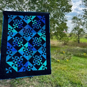 May include: A rectangular quilt featuring a repeating geometric pattern of pinwheel blocks in shades of blue, teal, and cyan, set against a solid black background with a black border featuring a continuous swirling quilted stitch pattern.