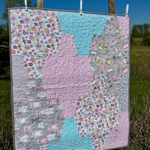 May include: A handmade quilt with a patchwork design, featuring hexagonal shapes in pink, light blue, and gray. The quilt showcases floral and animal patterns, with a gray border and intricate stitching. It is hanging outdoors on a sunny day.
