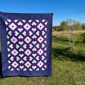 May include: A handmade quilt with a geometric pattern. The quilt features a central design of white and purple shapes against a purple background, framed by a dark blue border. The quilt is displayed outdoors on a sunny day.