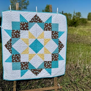 May include: A square quilt with a star pattern, displayed outdoors. The quilt features a white background with a teal, brown, and yellow star design. The quilt is held up by clothespins on a wooden frame, with a green field and blue sky in the background.