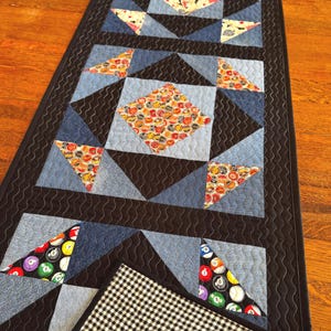 May include: A rectangular quilted table runner featuring a patchwork design of denim, black fabric, and patterned cotton squares. The pattern includes billiard ball motifs and circular graphics. The runner has a black border and a black and white gingham backing.