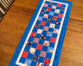 May include: A blue and white table runner with a red, white, and blue checkered pattern. The runner is made of fabric and has a white border.