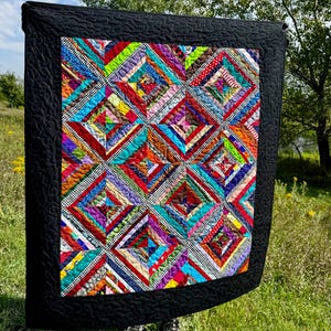 May include: A rectangular quilt featuring a central panel of multi-colored fabric strips arranged in a diamond pattern, surrounded by a solid black border with a swirling quilted texture. The fabric strips include various colors and patterns.