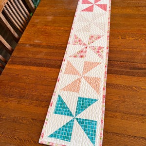 May include: A quilted table runner featuring a pinwheel design. The runner showcases alternating pinwheels in shades of pink, coral, and teal, set against a cream background. The edges are finished with a floral print border, adding a touch of color.