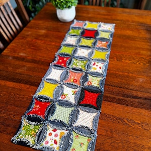 May include: A denim table runner with a patchwork design. The runner features square patches in various colors and floral patterns, including red, yellow, and green. The edges are frayed, giving it a rustic look. The runner is placed on a dark wooden table.