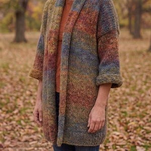 Puede incluir: Un cárdigan abierto de punto grueso con una paleta de colores variados en tonos tierra apagados, incluyendo óxido, verde oliva y azul pizarra. La prenda presenta un cuello chal ancho, hombros caídos y mangas tres cuartos con puños.