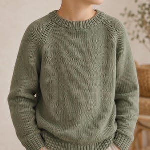 May include: A sage green knit sweater with a ribbed crew neck and cuffs. The long-sleeved pullover has a classic design and a relaxed fit. The sweater is made of a soft, textured material, perfect for cooler weather. The sweater is a children's clothing item.