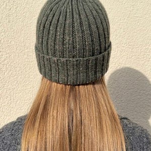 Könnte beinhalten: Eine dunkel olivgrüne Strickmütze mit gerippter Textur und umgeschlagenem Saum. Die Mütze ist mit einem vertikalen Strickmuster gefertigt, das von der Krone bis zum Rand verläuft und durchgehend aus einheitlichem dunkelgrünem Garn besteht.
