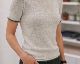 Può includere: Un maglione in maglia a maniche corte di colore beige mélange chiaro. Il capo presenta uno scollo rotondo, maniche corte e un orlo dritto, ciascuno rifinito con un dettaglio di bordo a contrasto in verde bosco scuro.