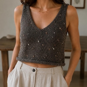 May include: A dark charcoal grey sleeveless knit tank top featuring a V-neckline and a speckled texture with small flecks of white and tan throughout. The garment has a thick ribbed hem and is paired with light beige trousers with a button closure.