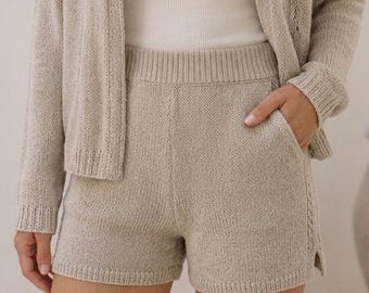Può includere: Pantaloncini in maglia beige con vita alta elasticizzata, tasche laterali e un piccolo spacco laterale sull'orlo. I pantaloncini sono realizzati in tessuto a maglia testurizzato e sono abbinati a un cardigan aperto in maglia coordinato.