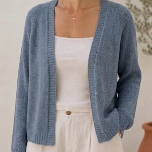 Peut inclure: Un cardigan en tricot bleu poussiéreux ouvert sur le devant, doté de manches longues avec poignets côtelés, d'un ourlet côtelé et d'une patte de boutonnage côtelée. Il est porté sur un débardeur blanc côtelé et un pantalon clair à bouton marron.