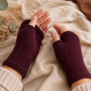 May include: A pair of deep burgundy fingerless gloves featuring a smooth stockinette stitch pattern on the main body and a textured garter stitch border at the wrist and finger openings. The gloves cover the palm and wrist area while leaving fingers exposed.