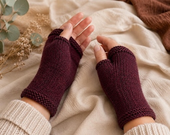 Può includere: Un paio di guanti senza dita color bordeaux scuro caratterizzati da un motivo a maglia rasata sul corpo principale e un bordo testurizzato a legaccio sul polso e sulle aperture delle dita, che coprono il palmo e il polso.