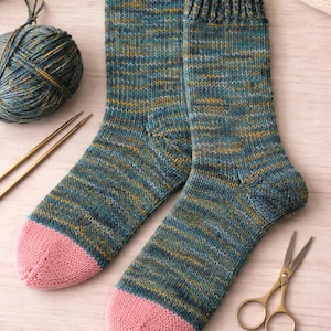 Può includere: Un paio di calze lavorate a maglia a mano con un motivo in filato variegato color ottanio, blu e giallo, con punte rosa a tinta unita e polsini a coste. Le calze sono presentate insieme a un gomitolo abbinato, due ferri e un paio di forbici.
