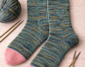 Può includere: Un paio di calze lavorate a maglia a mano con un motivo in filato variegato color ottanio, blu e giallo, con punte rosa a tinta unita e polsini a coste. Le calze sono presentate insieme a un gomitolo abbinato, due ferri e un paio di forbici.