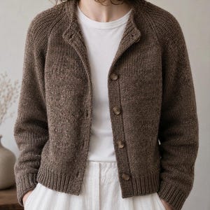 May include: A brown knit cardigan featuring a round neckline, long sleeves, and a front button closure with five matching brown buttons. The garment is crafted from a textured, heathered yarn and includes ribbed cuffs and a ribbed hemline.