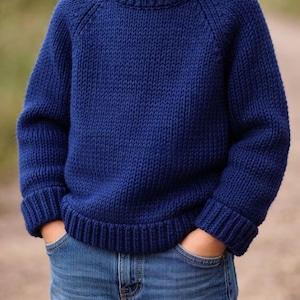May include: A solid navy blue long-sleeve knit sweater featuring a thick ribbed crew neckline, ribbed cuffs, and a ribbed hem. The sweater is paired with blue denim jeans that have visible pockets where hands are placed inside.