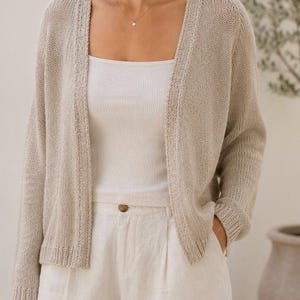 Puede incluir: Un cárdigan de punto beige con mangas largas, usado sobre una camiseta sin mangas blanca y combinado con pantalones color crema. El cárdigan tiene un diseño sencillo y abierto. El conjunto se completa con un delicado collar dorado.