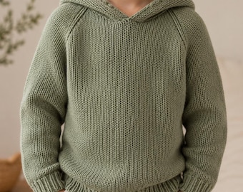 Può includere: Un maglione con cappuccio color verde salvia a maniche lunghe, caratterizzato da una trama a maglia grossa, maniche raglan e polsini a coste sui polsi e sull'orlo. Il capo presenta un motivo a maglia visibile e un cappuccio sul collo.