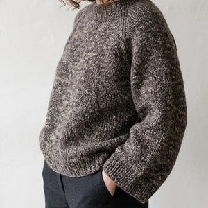 May include: A long-sleeved, crew-neck sweater crafted from a thick, heathered yarn in shades of dark brown, charcoal, and cream. The garment features a ribbed knit collar, cuffs, and hem, with a relaxed fit and raglan sleeves in a chunky knit texture.