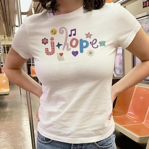 May include: A white short-sleeved t-shirt featuring the text j-hope in colorful, patterned letters. The design includes various small graphic icons such as a winking emoji, a music note, a star, a flower, a butterfly, and a heart with the word love.