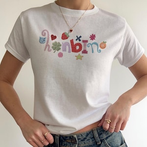 May include: A white short-sleeved t-shirt featuring a colourful graphic print that spells out the word "hanbin" in various patterned letters, accompanied by small icons including a strawberry, a clover, a star, and a heart.