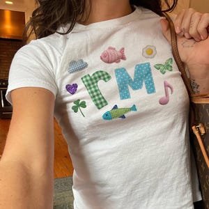 May include: A white short-sleeved t-shirt featuring the letters rM in a green gingham and blue polka dot pattern. The shirt is decorated with various small graphics including a pink fish, fried egg, butterfly, purple heart, clover, and music note.